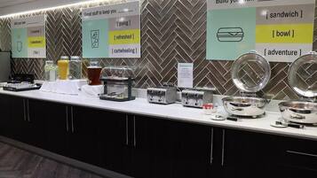 A buffet setup with various food items and utensils, including toasters and bowls, against a herringbone patterned wall.