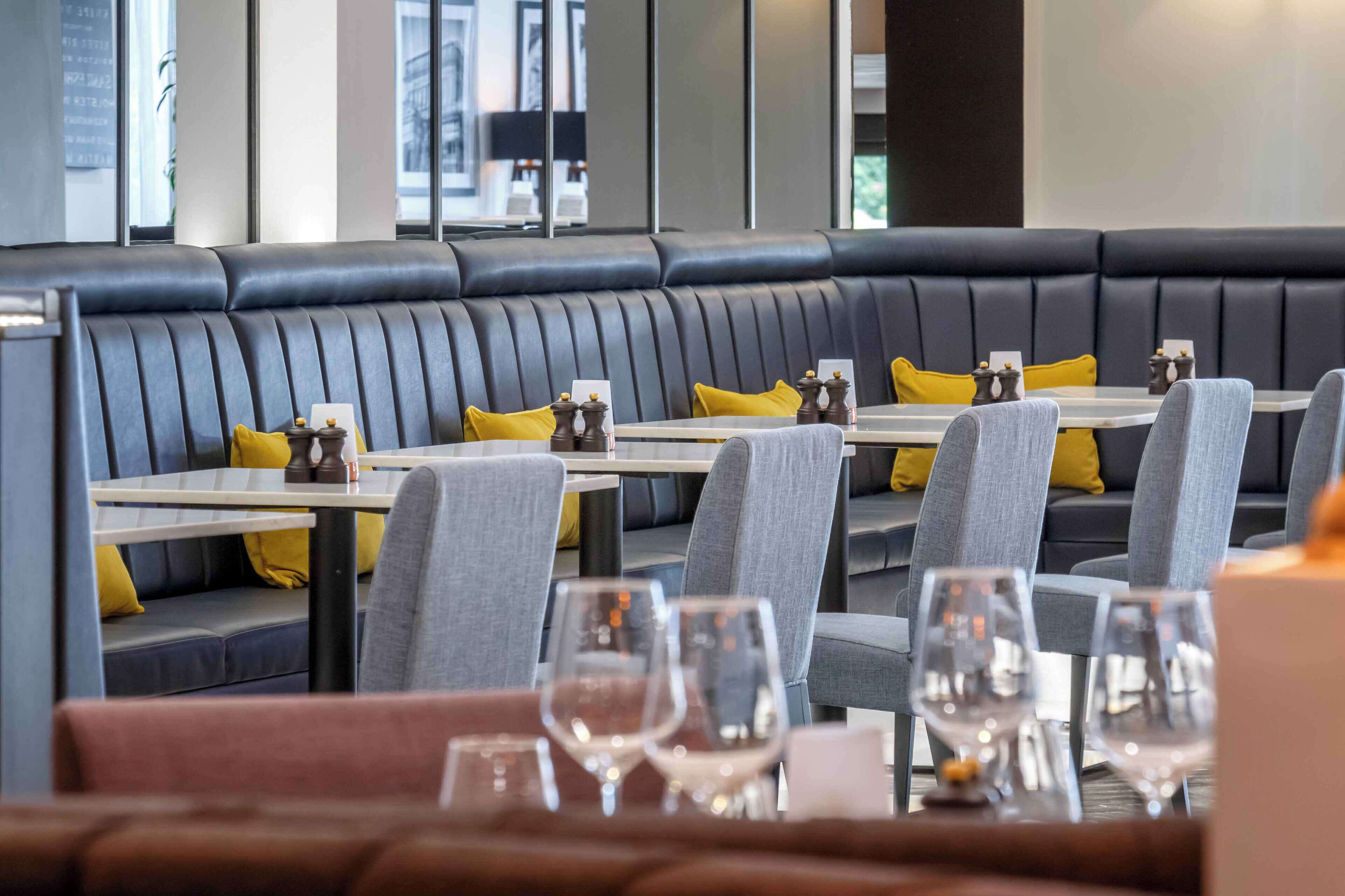 Un ristorante moderno con una lunga panca in pelle nera, sedie rivestite di grigio e cuscini gialli. I tavoli sono apparecchiati con bicchieri da vino e portaposate.