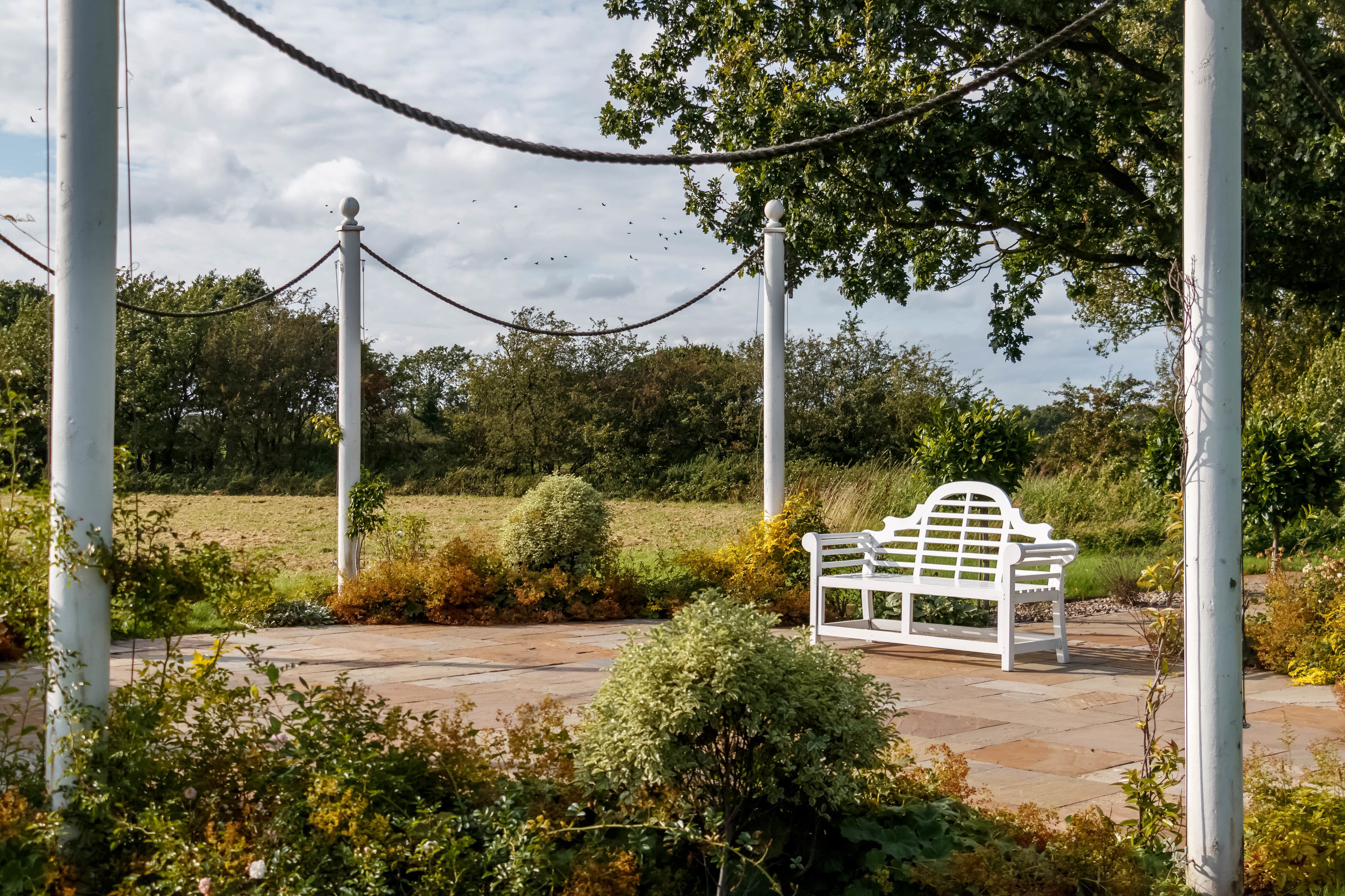 Un banc blanc dans un jardin, délimité par une barrière faite de cordes et agrémenté de colonnes blanches.