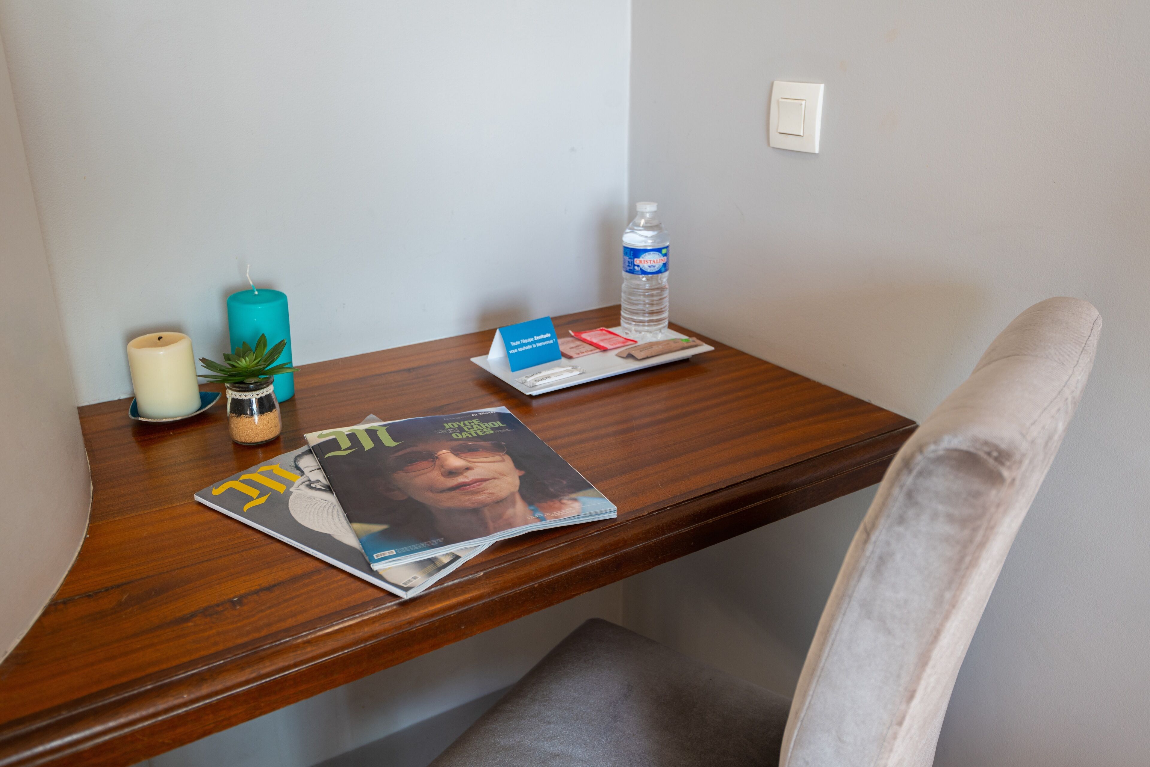 A wooden desk with a magazine, a candle, a small plant, and a water bottle.