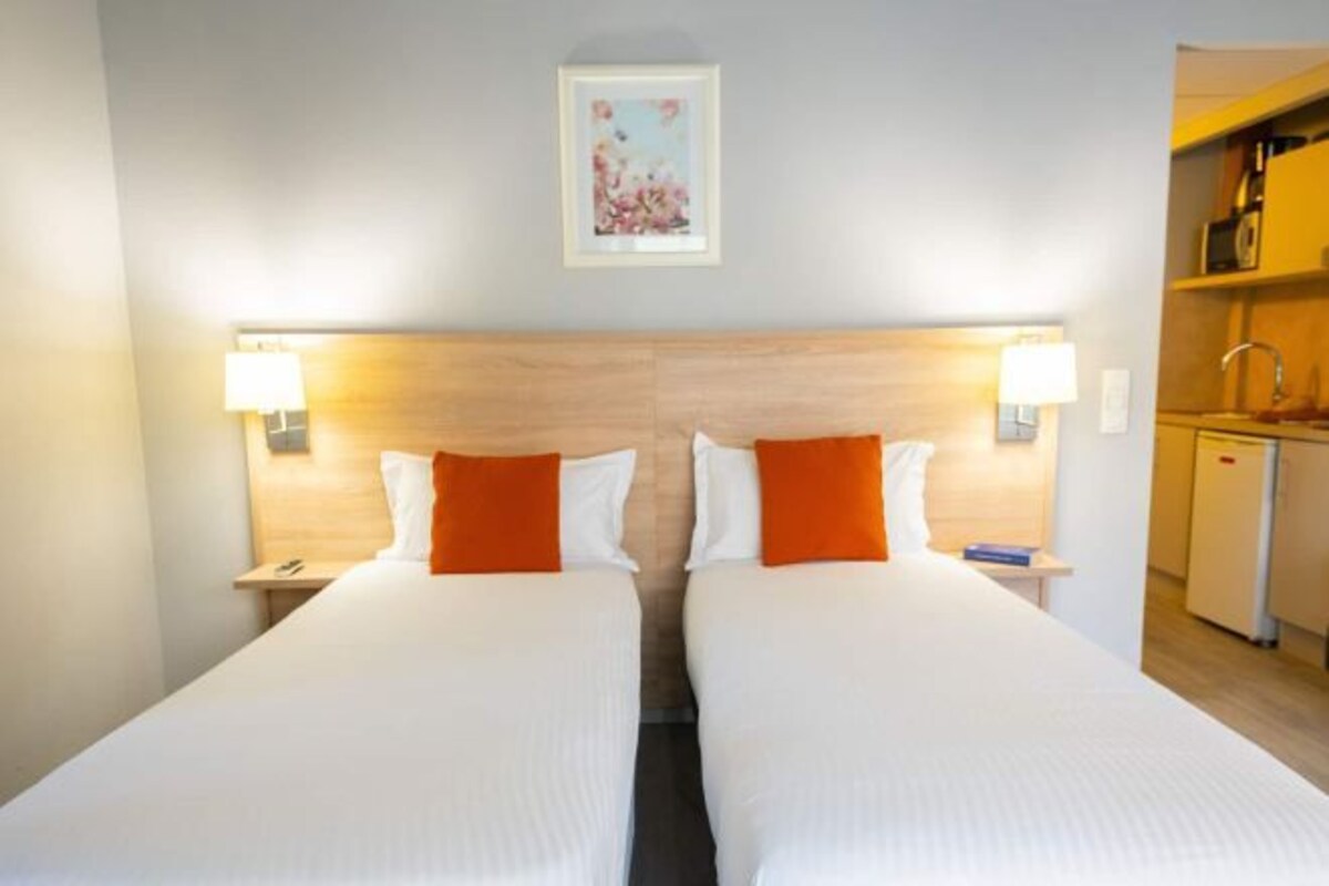 A double bed with white and orange bedding, two bedside lamps, and a small kitchenette.