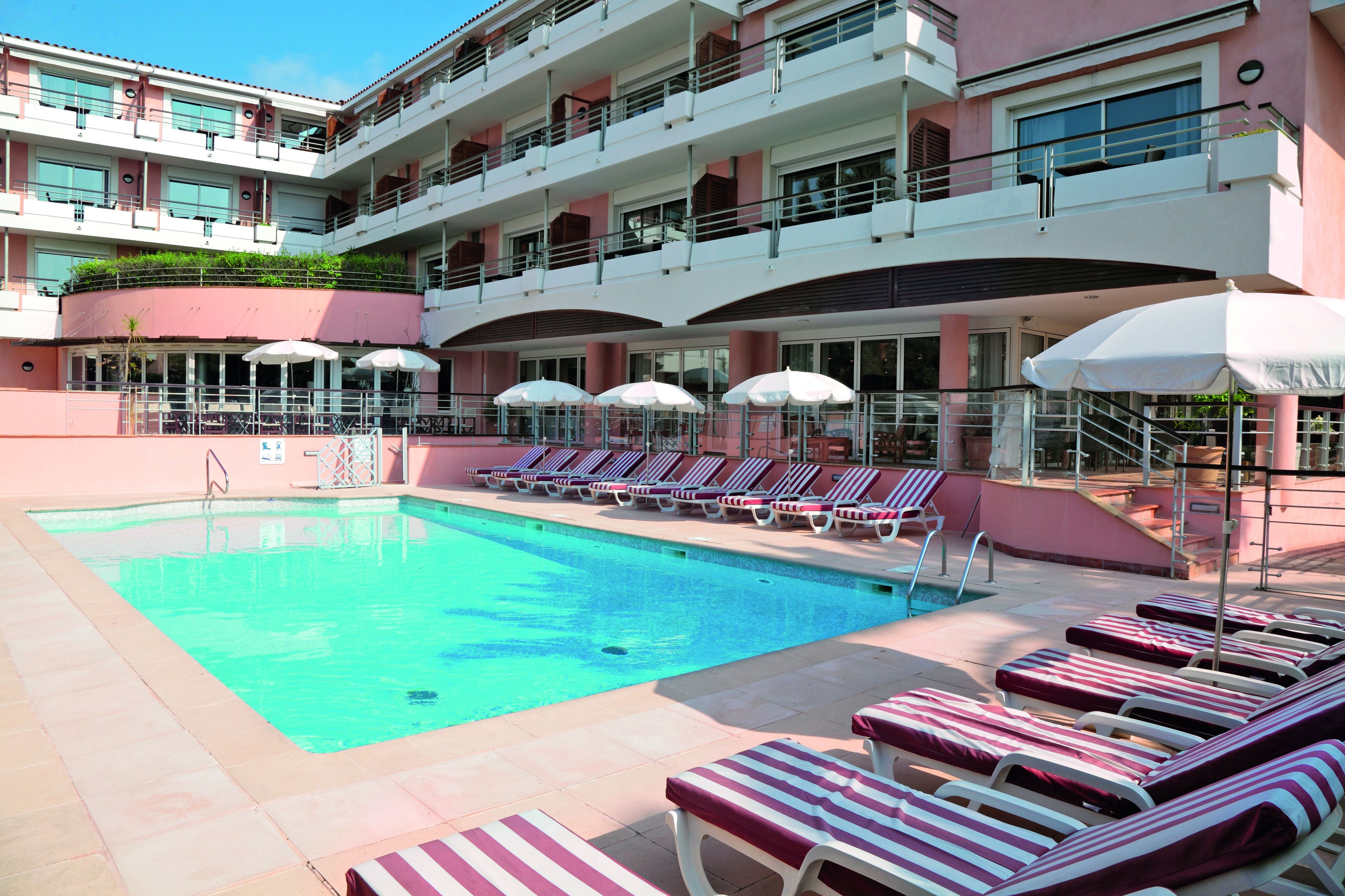 Zona de piscina de hotel con tumbonas y sombrillas, una piscina cristalina y un edificio de varios pisos con balcones.