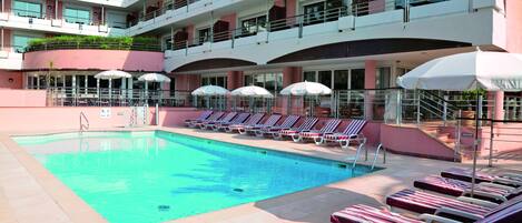 Zona de piscina de hotel con tumbonas y sombrillas, una piscina cristalina y un edificio de varios pisos con balcones.