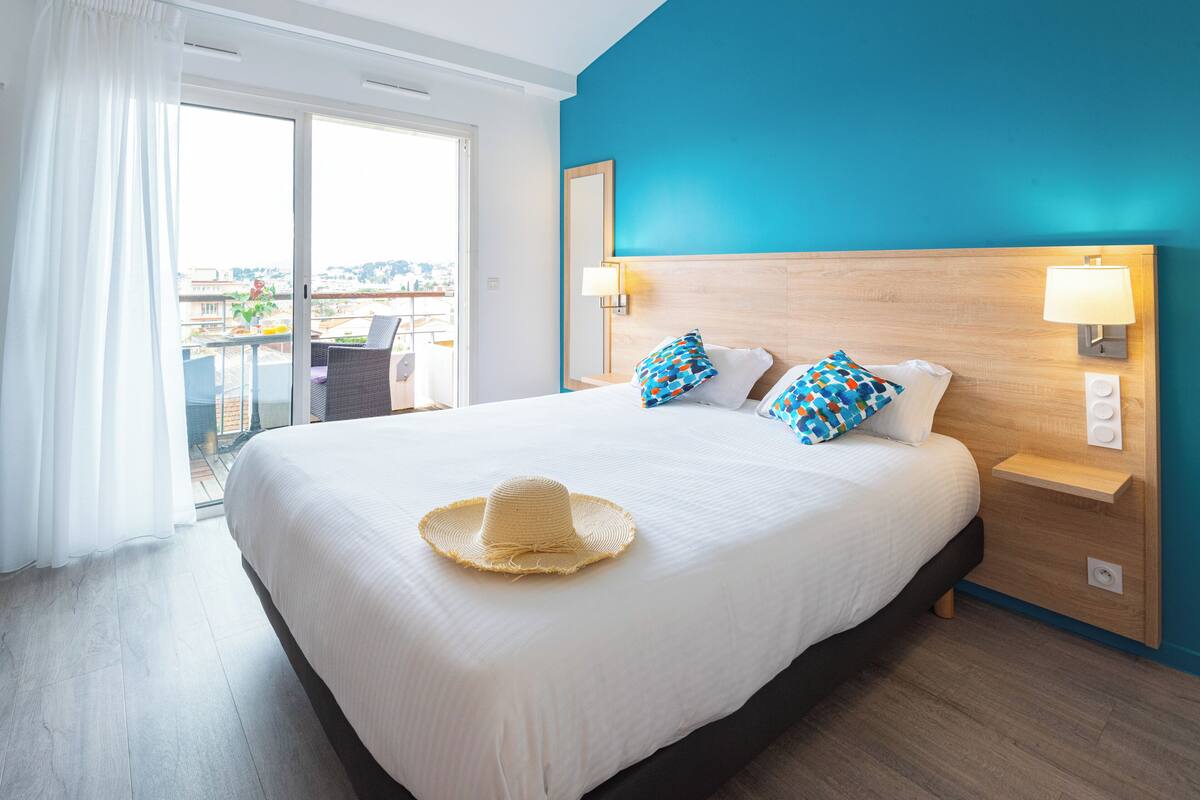 A hotel room with a large bed, two pillows, a nightstand, a lamp, and a view of the city through a sliding door.