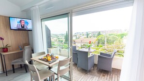 Habitación de hotel moderna con balcón, zona de comedor con mesa y sillas, televisor montado en la pared y vistas a la ciudad.
