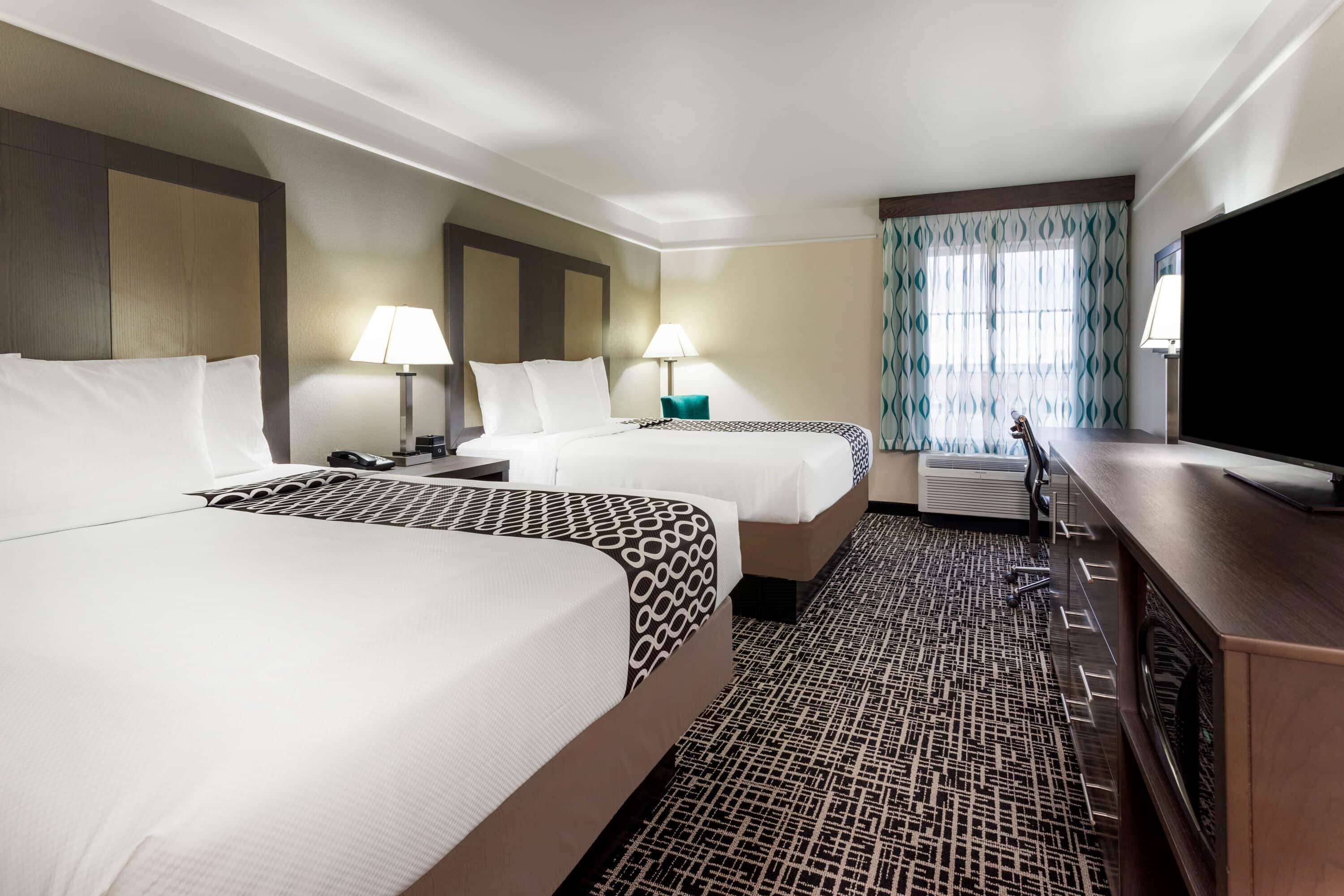 A hotel room with two beds, a TV, a desk, and a window with curtains.