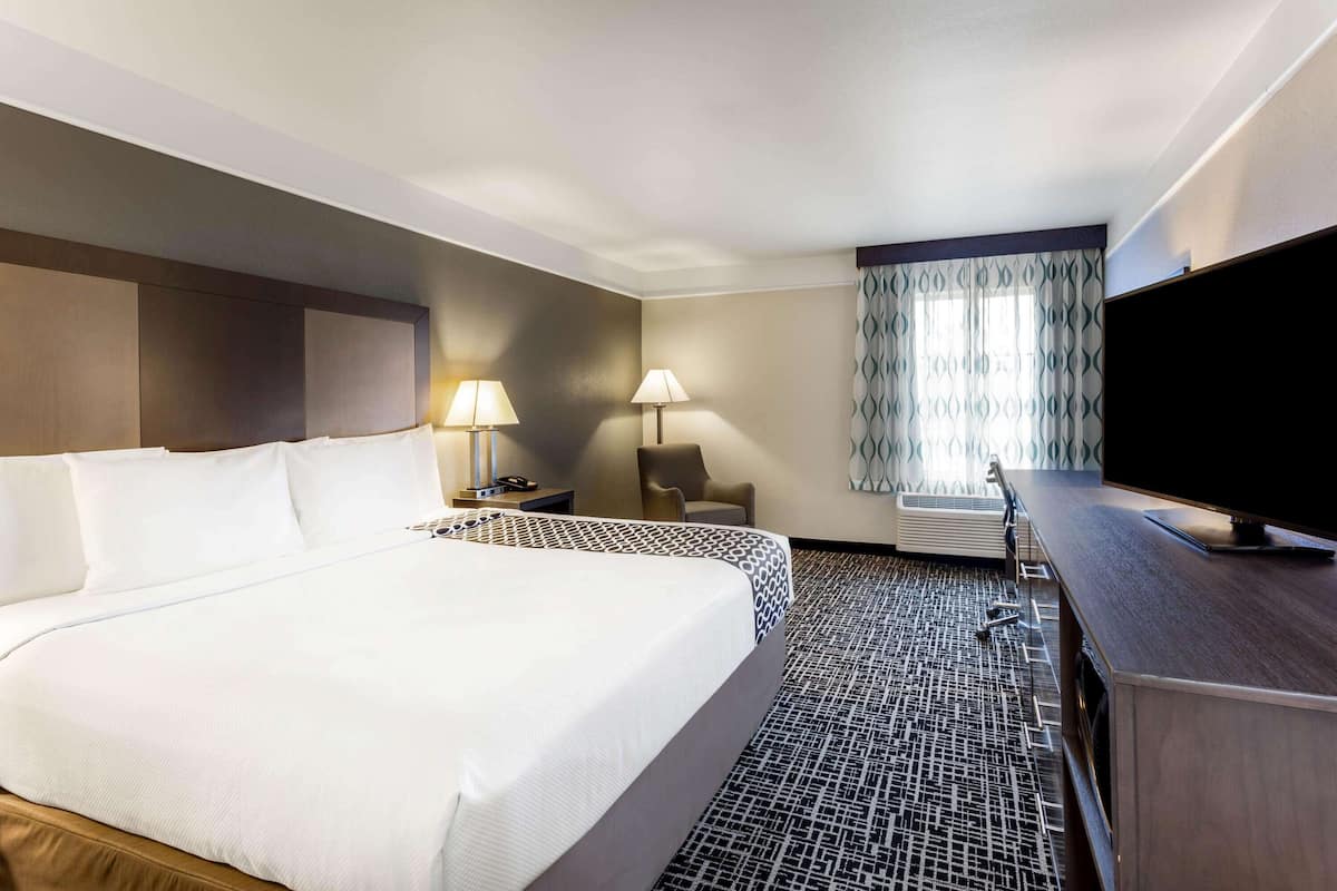 Une chambre d’hôtel avec un grand lit, deux tables de chevet avec des lampes, un téléviseur à écran plat, un fauteuil et une fenêtre agrémentée de rideaux à motifs.