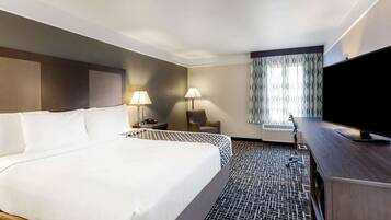 Une chambre d’hôtel avec un grand lit, deux tables de chevet avec des lampes, un téléviseur à écran plat, un fauteuil et une fenêtre agrémentée de rideaux à motifs.
