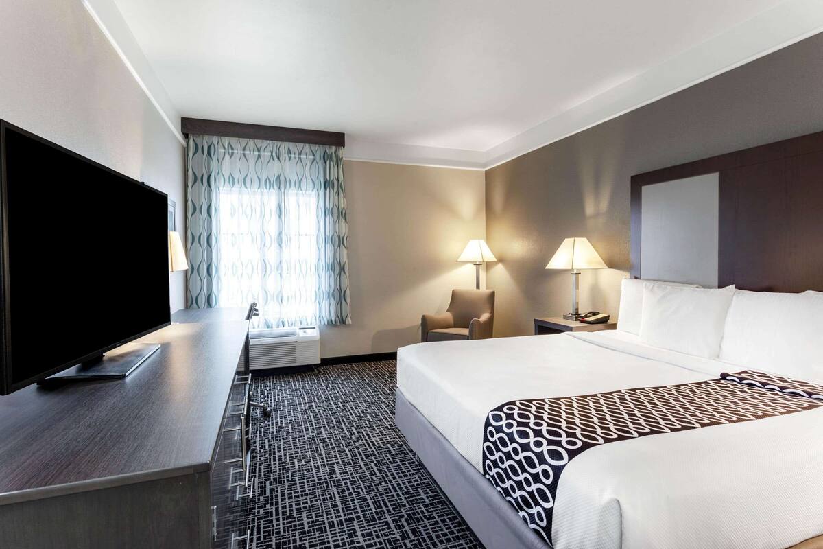 Une chambre d’hôtel comprenant un lit, un bureau avec un téléviseur à écran plat, deux lampes de chevet, une chaise et une grande fenêtre agrémentée de rideaux à motifs.