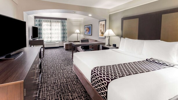 A hotel room with a large bed, a desk with a flat-screen TV, a seating area with a sofa and chairs, and a large window with curtains.