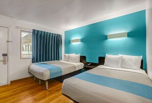 Quarto de hotel com duas camas, uma janela com cortinas, piso de madeira e uma parede de destaque em tom verde-água.
