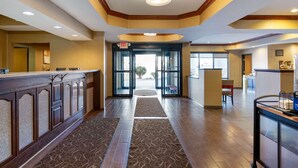 A hotel lobby with a reception desk, a glass door entrance, and a waiting area with a chair and a small table. - Comfort Suites South Haven near I-96 (South Haven)