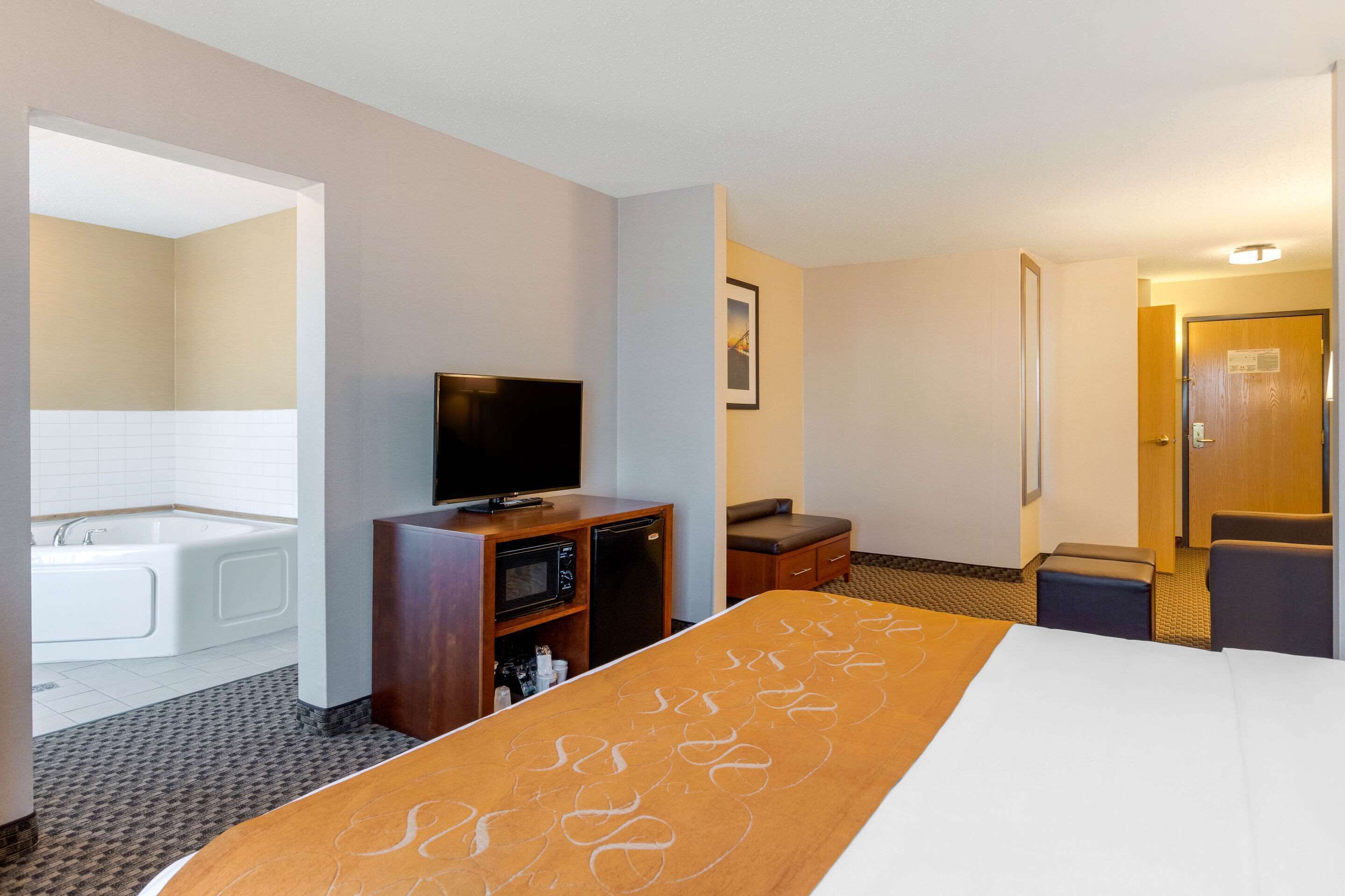 A hotel room with a bed, a television, a small desk, a microwave, a refrigerator, a bathroom with a bathtub, and a door leading to another room.