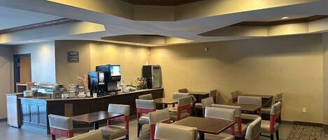 A hotel dining area with multiple tables and chairs, a counter with a coffee machine, and a patterned carpet.
