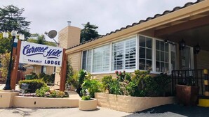 Un edificio de dos pisos con un letrero que dice "Carmel Ail Lodge" y un letrero de "Disponibilidad". Hay plantas en macetas y un pequeño jardín al frente.