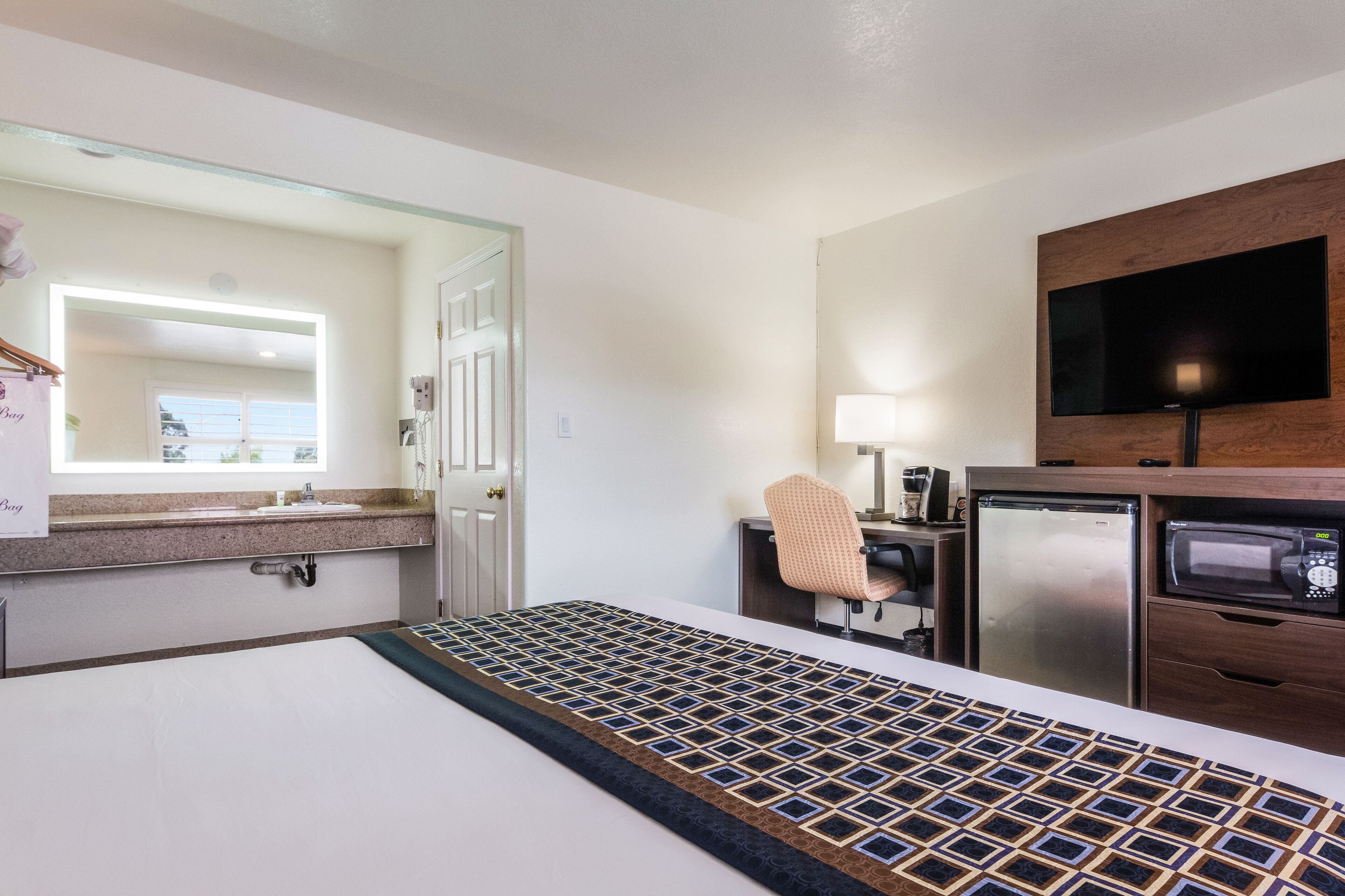 A hotel room with a bed, a desk, a refrigerator, a microwave, a television, and a bathroom with a sink and mirror.