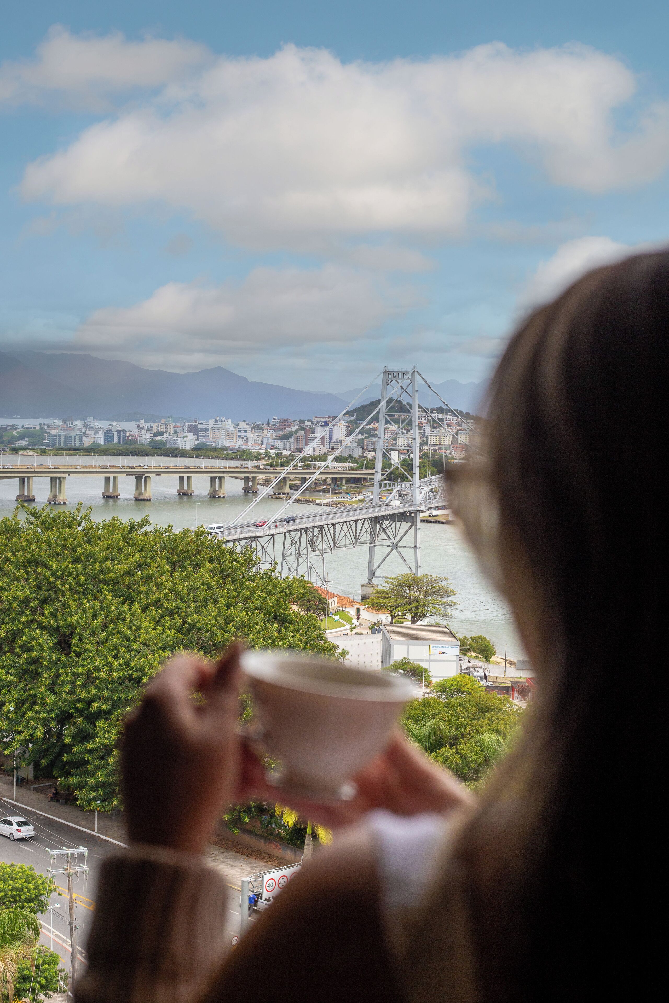 Eine Person hält eine Tasse in der Hand und blickt auf eine Brücke mit einer Stadtlandschaft im Hintergrund.
