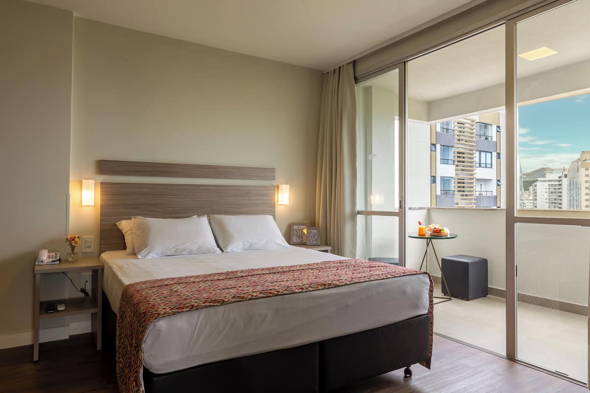 Une chambre d’hôtel moderne avec un grand lit, des tables de chevet, une petite table sur laquelle se trouve un panier de fruits, et une vue sur la ville grâce à de grandes fenêtres.