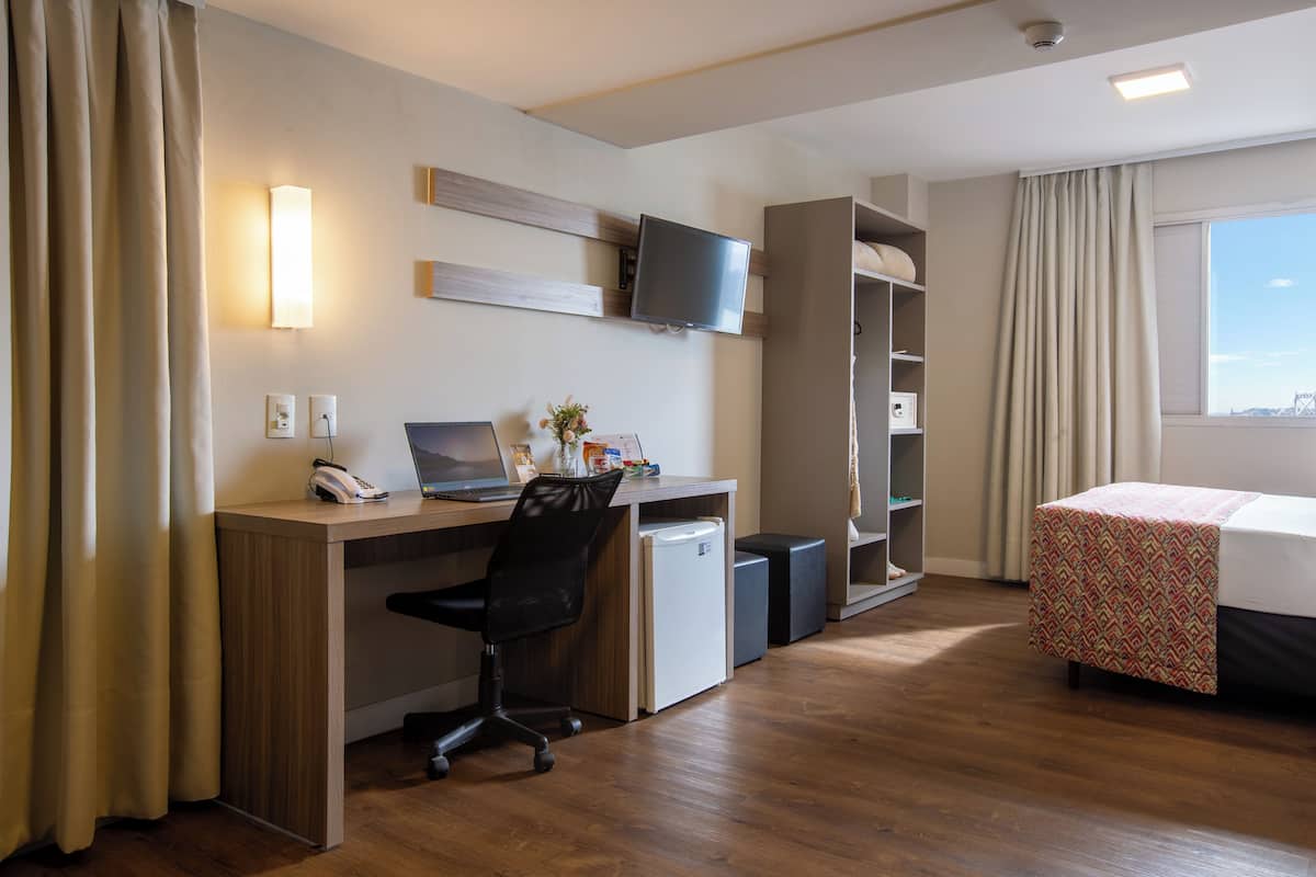 A hotel room with a wooden desk, a laptop, a small refrigerator, a bed with a patterned bedspread, a TV mounted on the wall, and a window with curtains.