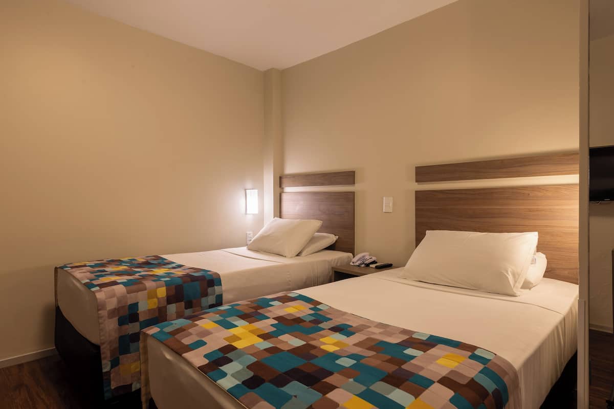Une chambre d’hôtel avec deux lits, chacun recouvert d’une couvre-lit à motifs différents, dotée d’une tête de lit en bois, d’une table de chevet avec une lampe et d’un téléphone fixé au mur.