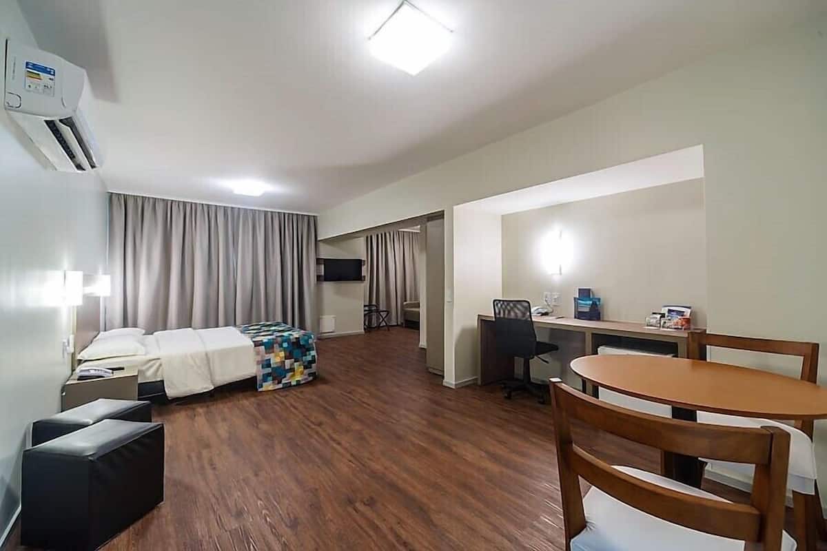A hotel room with a bed, a desk, a chair, a table, a TV, and an air conditioning unit.