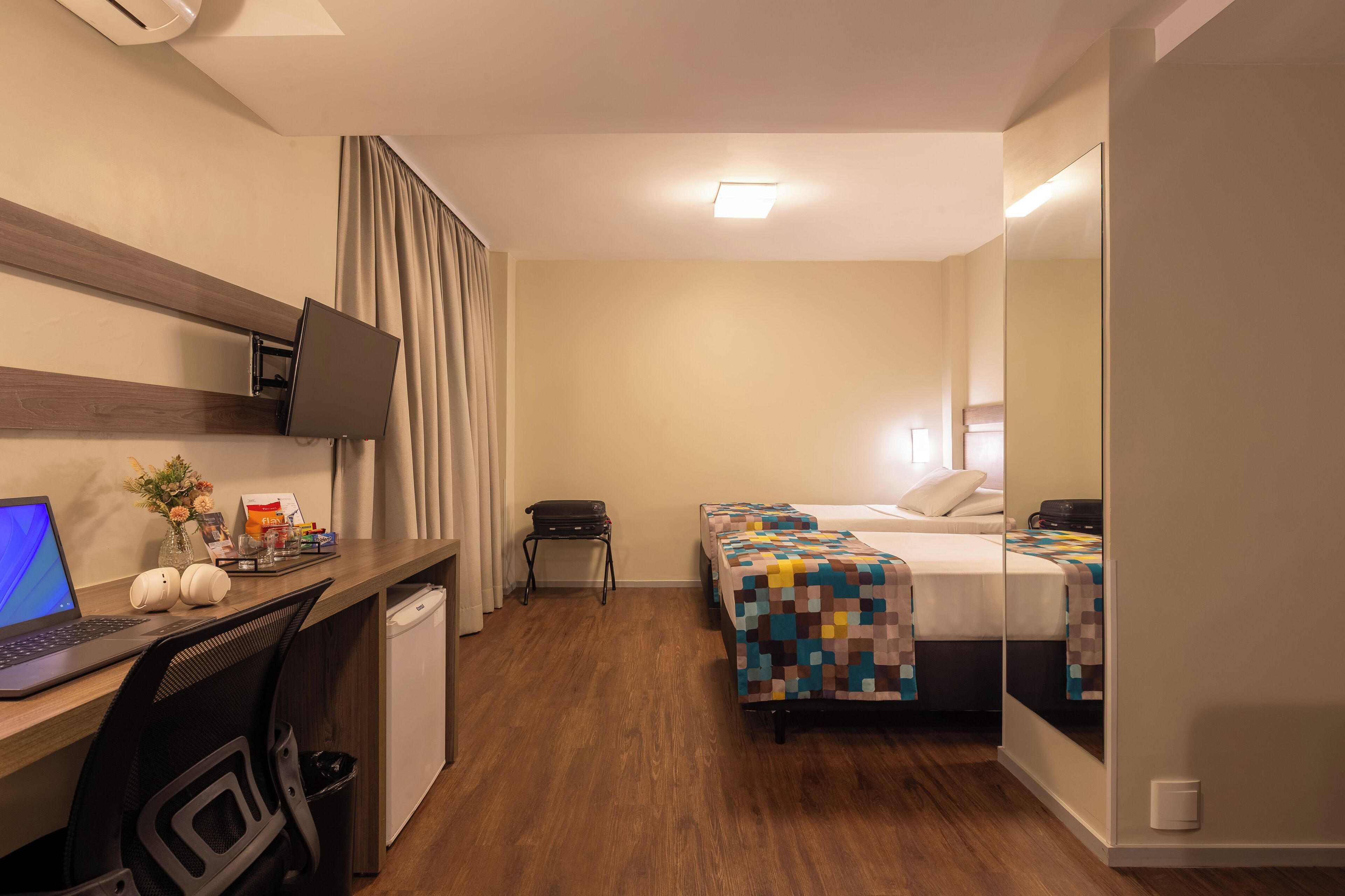 Habitación de hotel con escritorio, computadora portátil, dos camas, televisión y una pequeña mesa con un jarrón y flores.