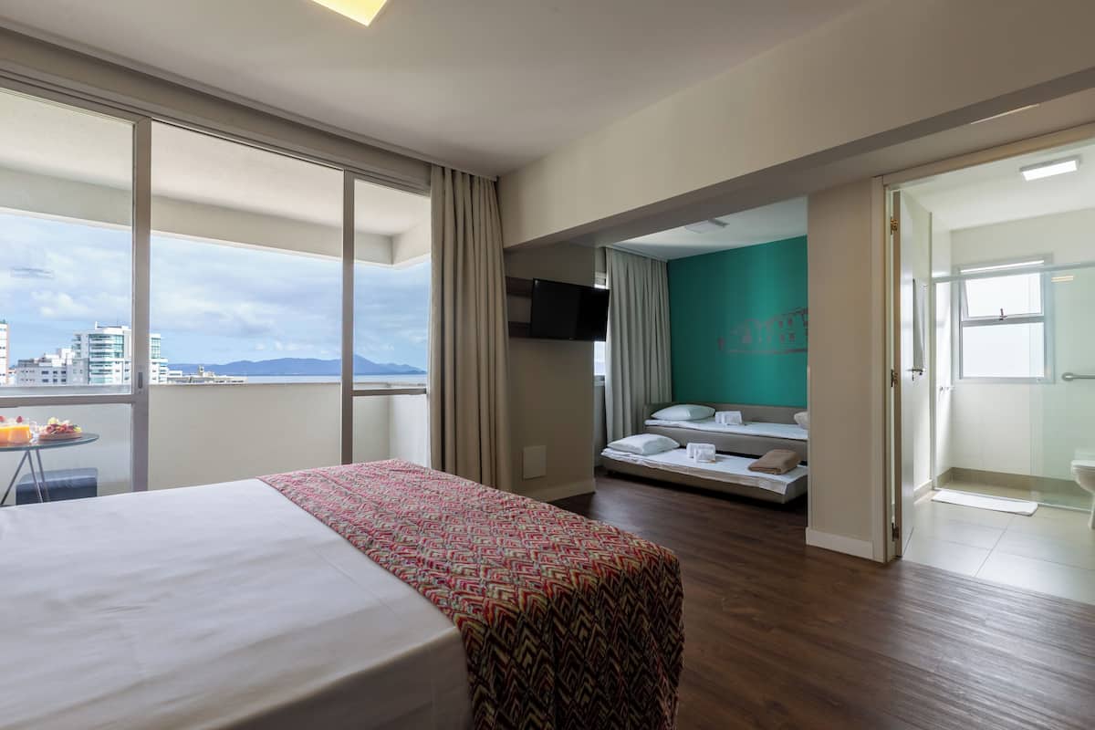 A hotel room with a large bed, a small seating area with a sofa and pillows, a TV mounted on the wall, and a balcony with a table and chairs.