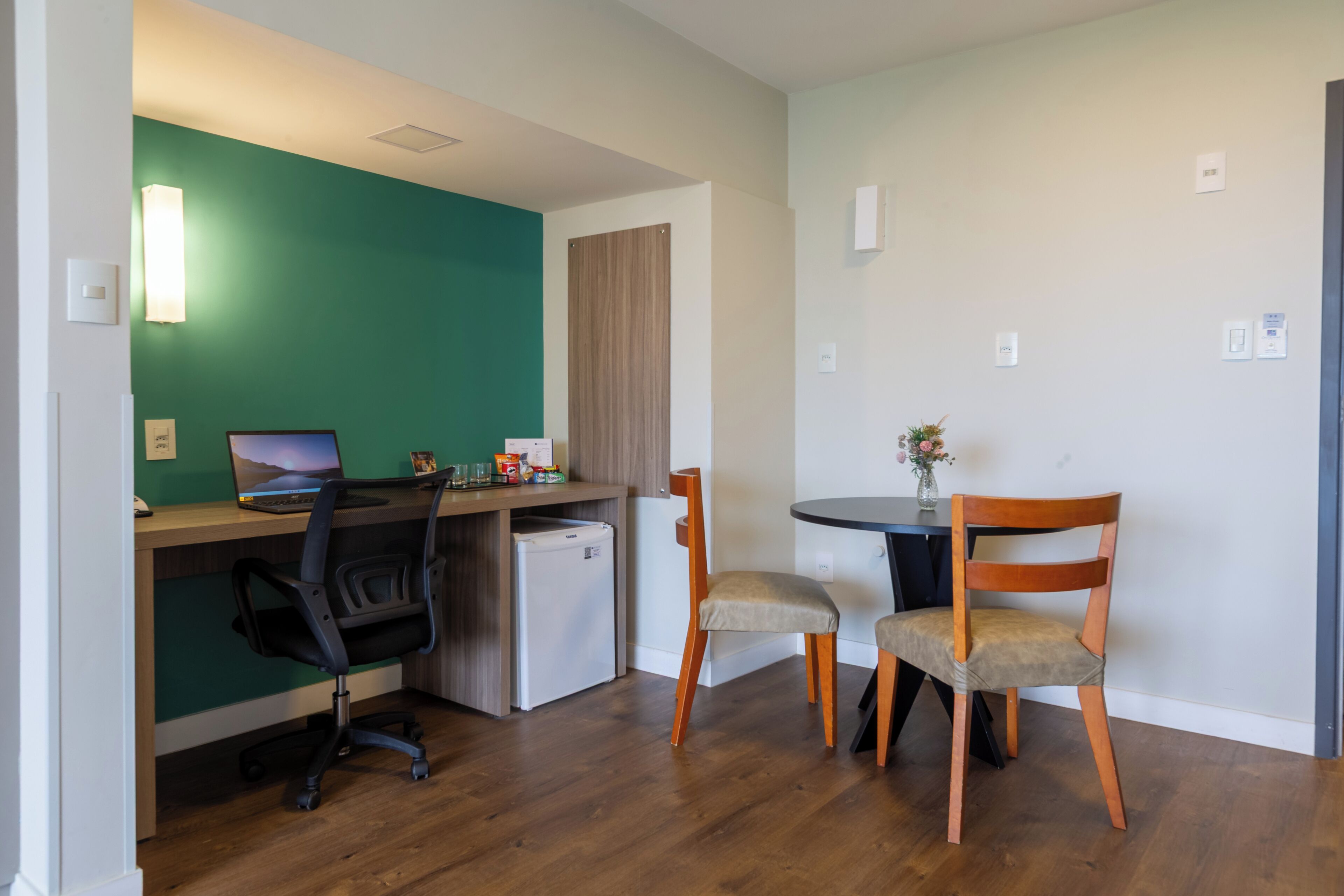 Un espacio habitable compacto con kitchenette, un escritorio con computadora portátil, dos sillas y un refrigerador pequeño.