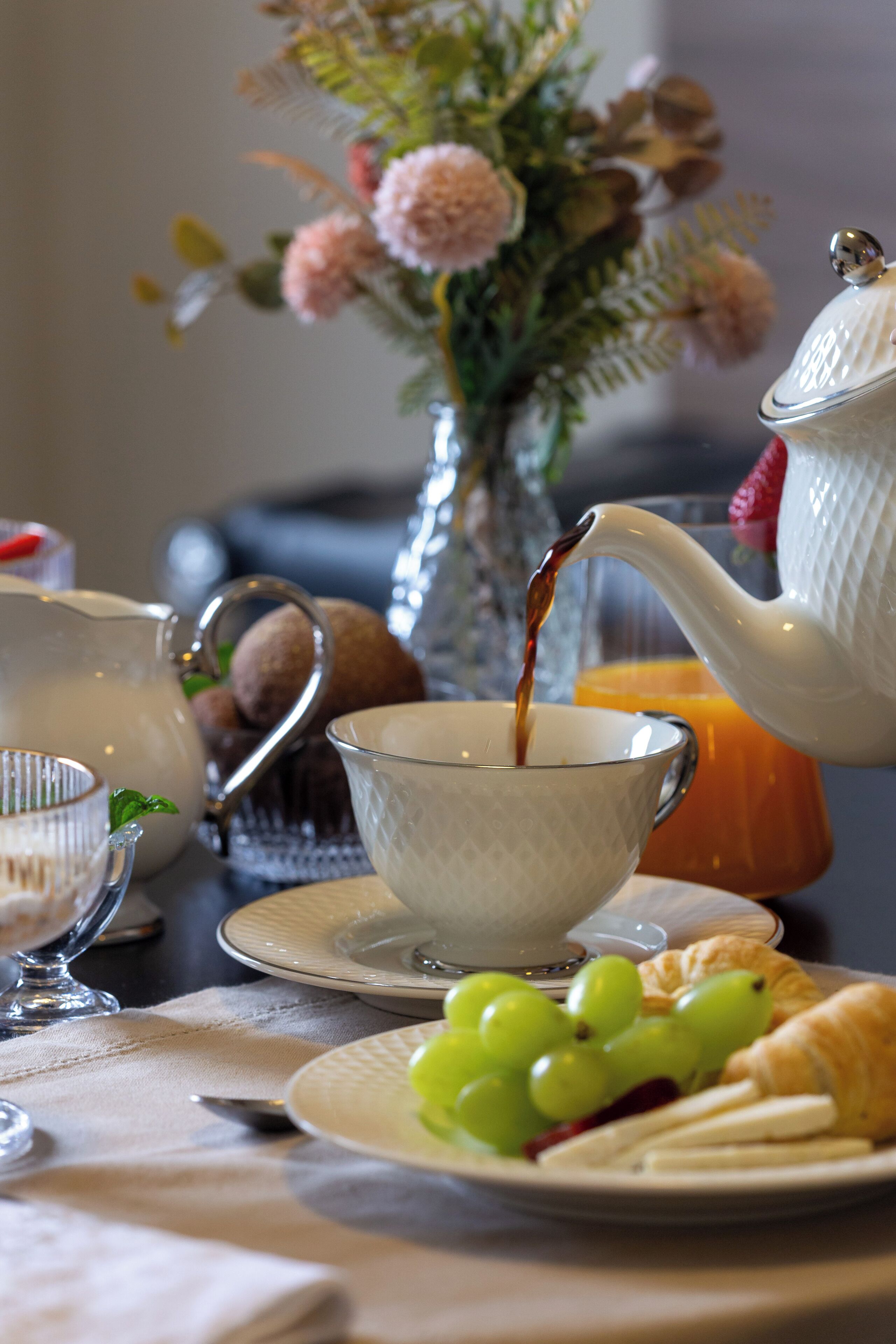 Uma mesa de café da manhã com um bule derramando chá em uma xícara, um copo de suco de laranja, um prato com uvas e um croissant, e um vaso com flores ao fundo.