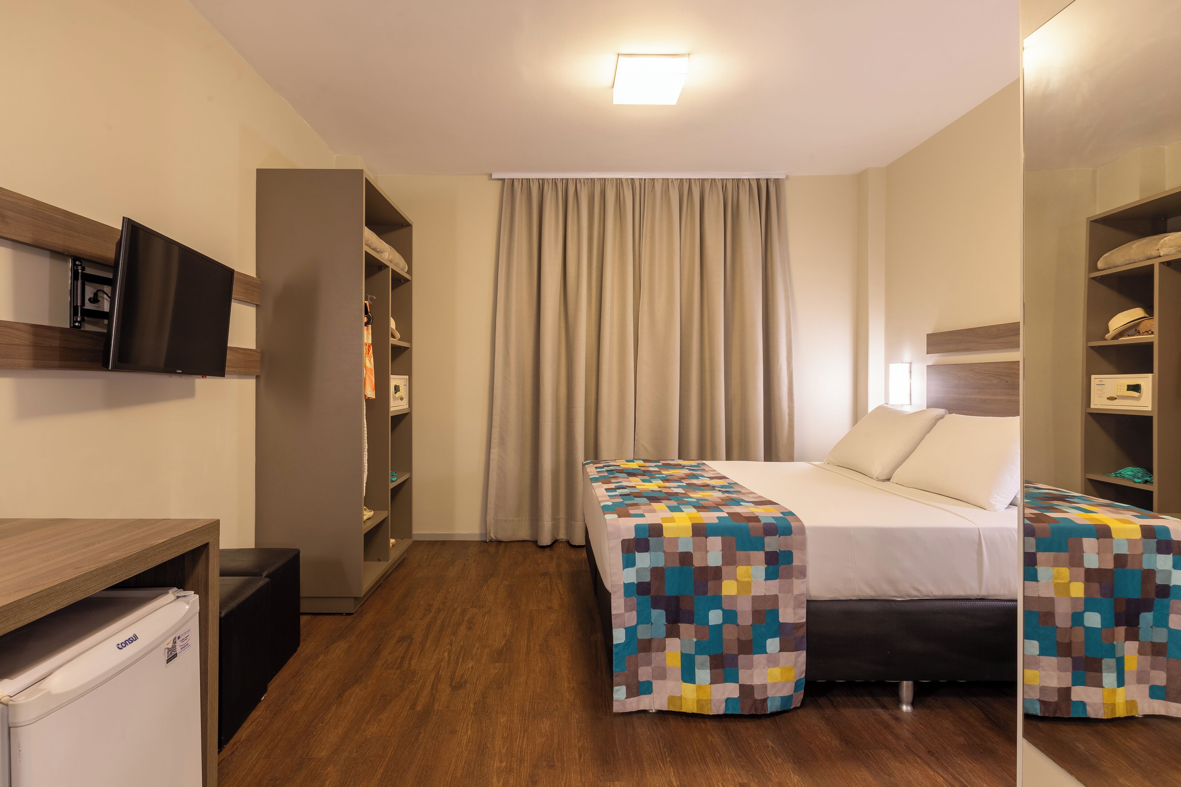 Quarto de hotel com cama, escrivaninha, guarda-roupa e televisão.