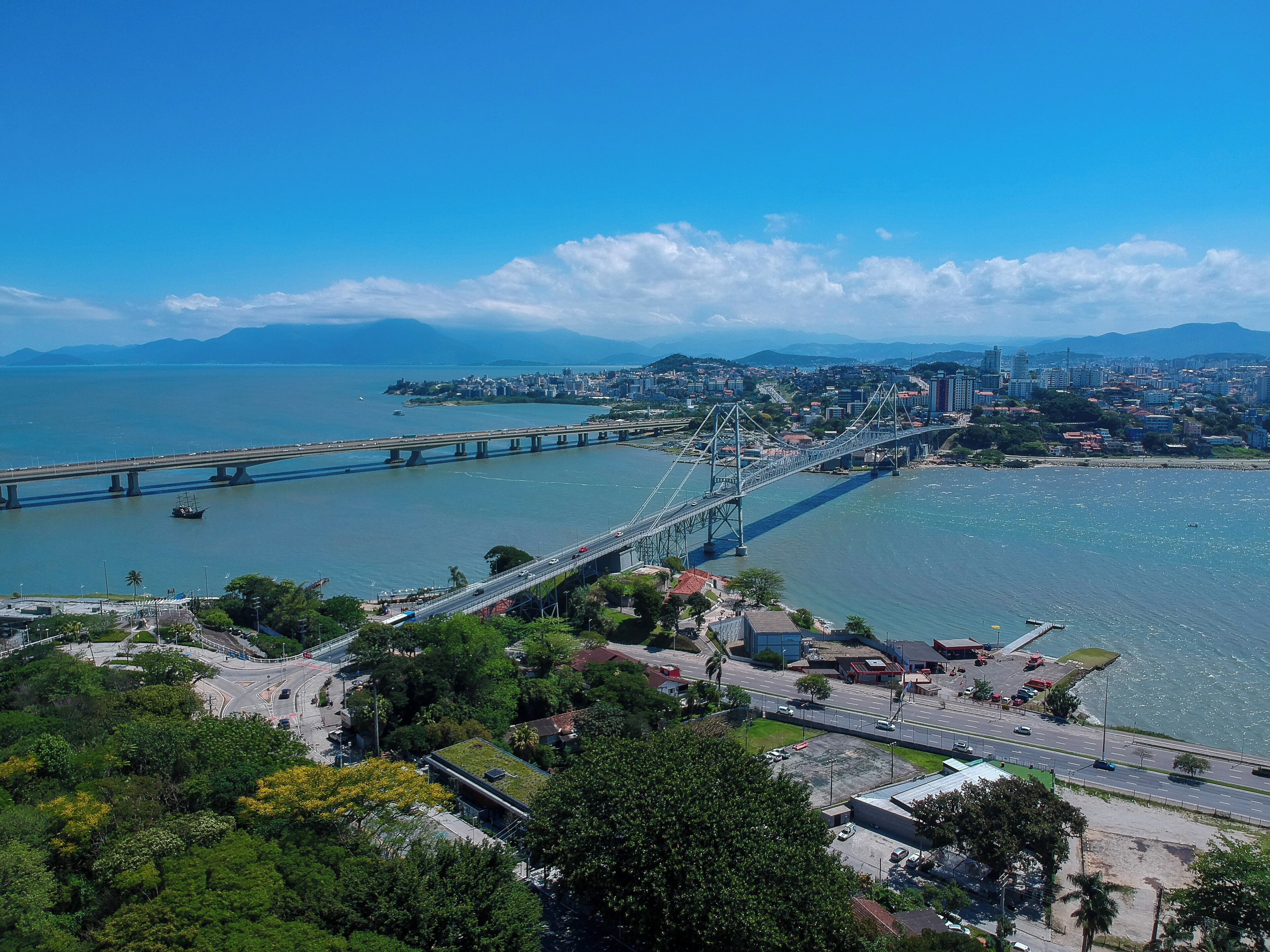 Un gran puente colgante cruza un río ancho, con un paisaje urbano y zonas verdes al frente.