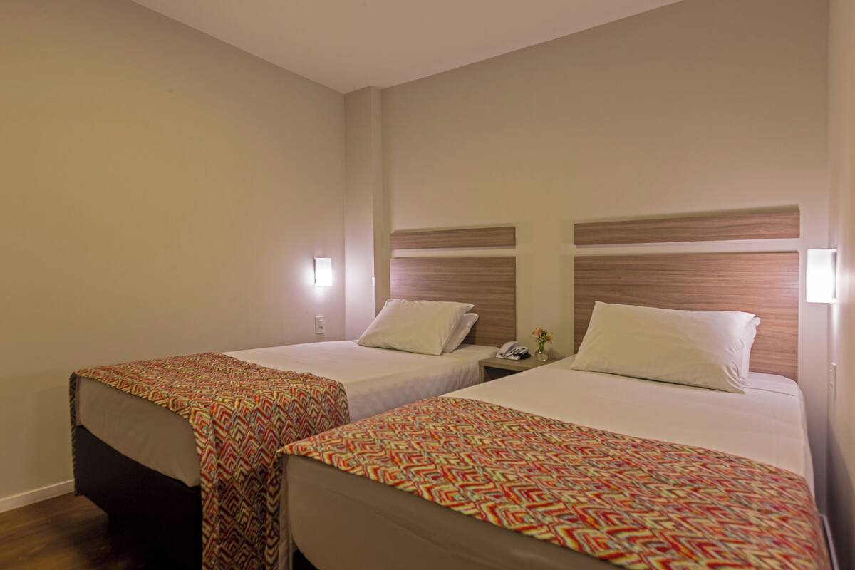 Une chambre d’h ôtel avec deux lits simples, chacun agrémenté d’une couvre-lit à motifs et d’une tête de lit en bois. On y trouve une table de chevet avec une lampe et un téléphone.