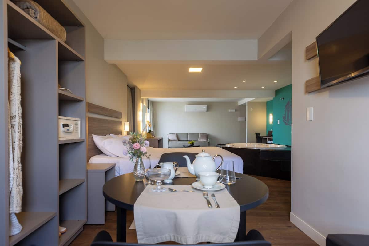 A modern hotel room with a dining area, a bed with a grey headboard, a small kitchen area, and a TV mounted on the wall.