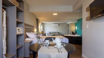 A modern hotel room with a dining area, a bed with a grey headboard, a small kitchen area, and a TV mounted on the wall.