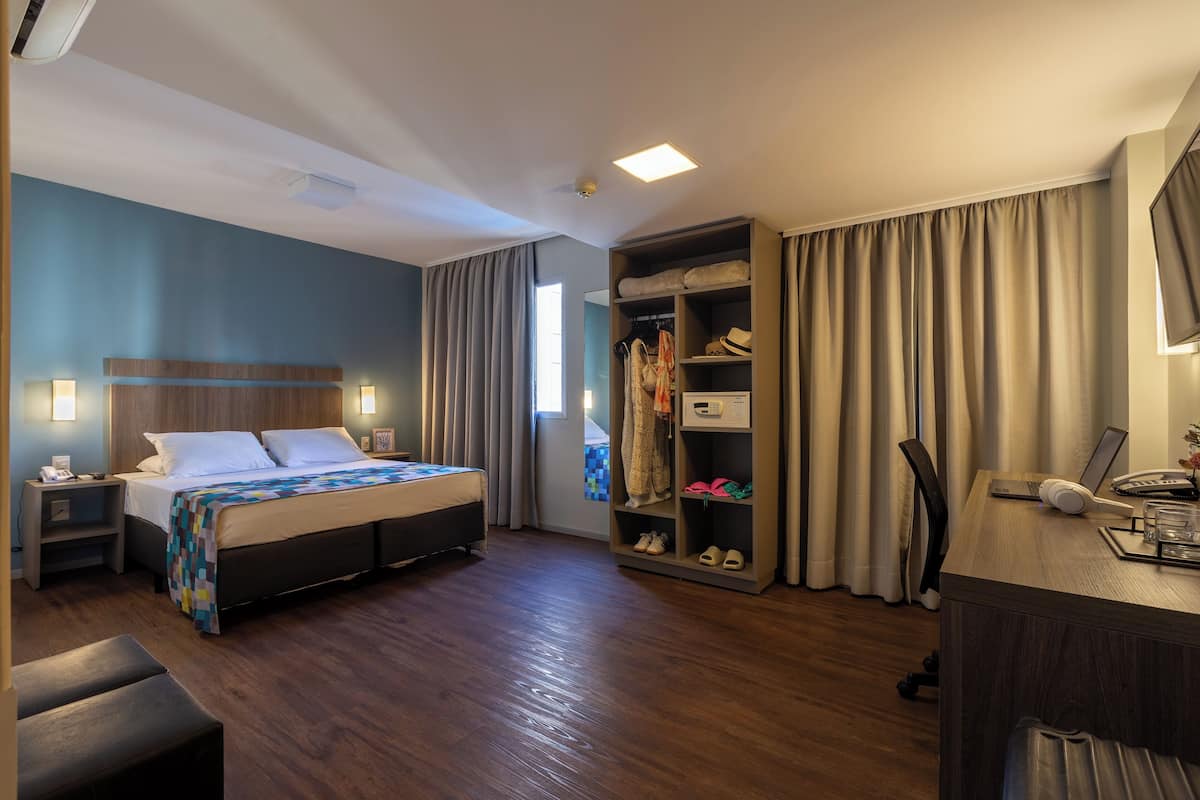 A hotel room with a bed, bedside tables, a desk with a computer, a wardrobe, and a large window with curtains.