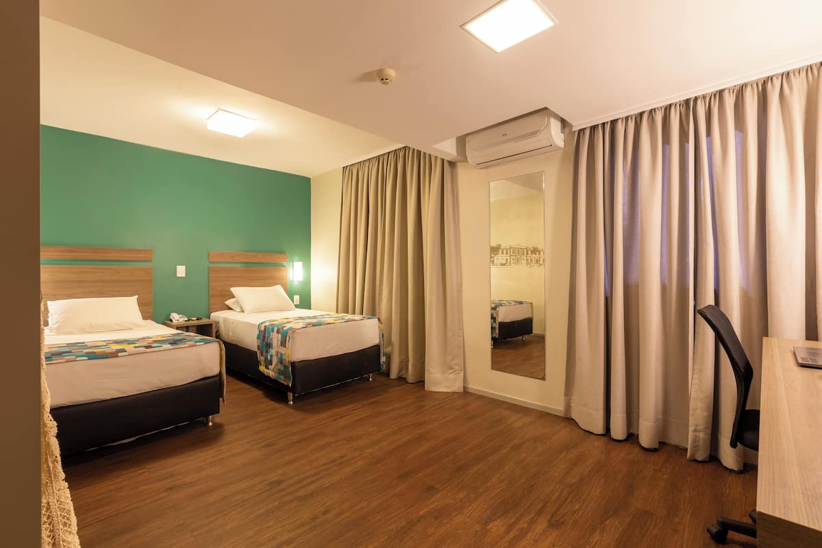 A hotel room with two beds, a desk, a chair, a mirror, and a window with curtains.