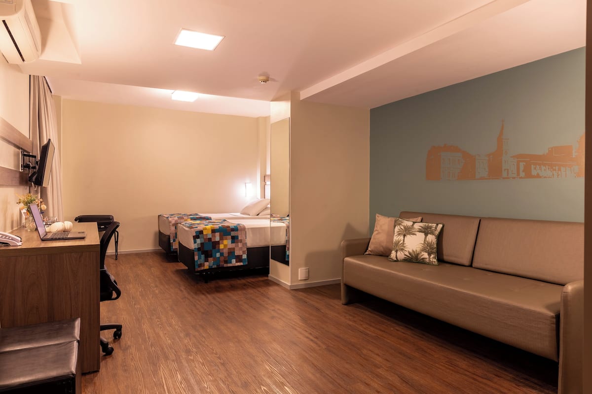 A modern hotel room with a wooden floor, a sofa, a desk with a laptop, and a bed with a colorful quilt.