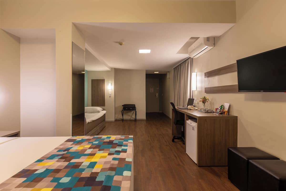 A hotel room with a bed, a desk, a chair, a television, and a small kitchenette.