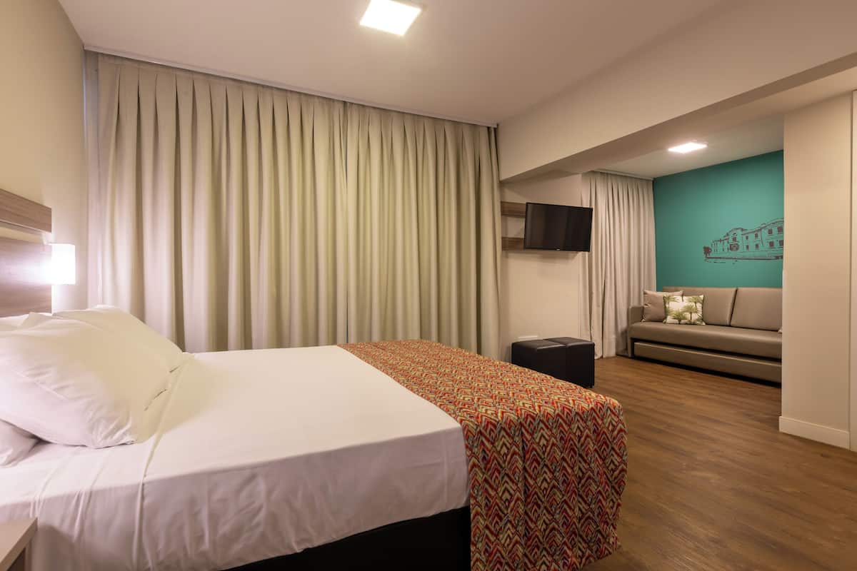 A hotel room with a bed, a sofa, a TV mounted on the wall, and a teal-colored accent wall.