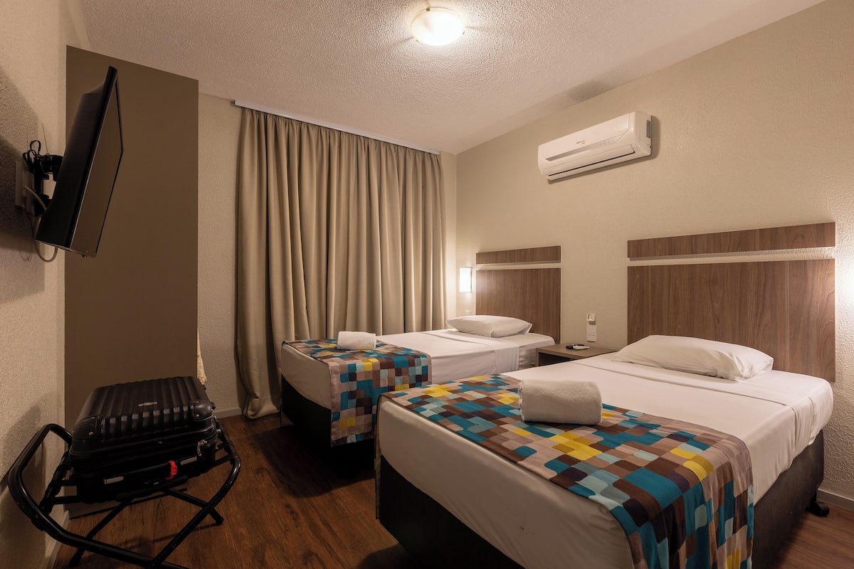 A hotel room with two beds, a desk, a chair, a suitcase, and an air conditioning unit.