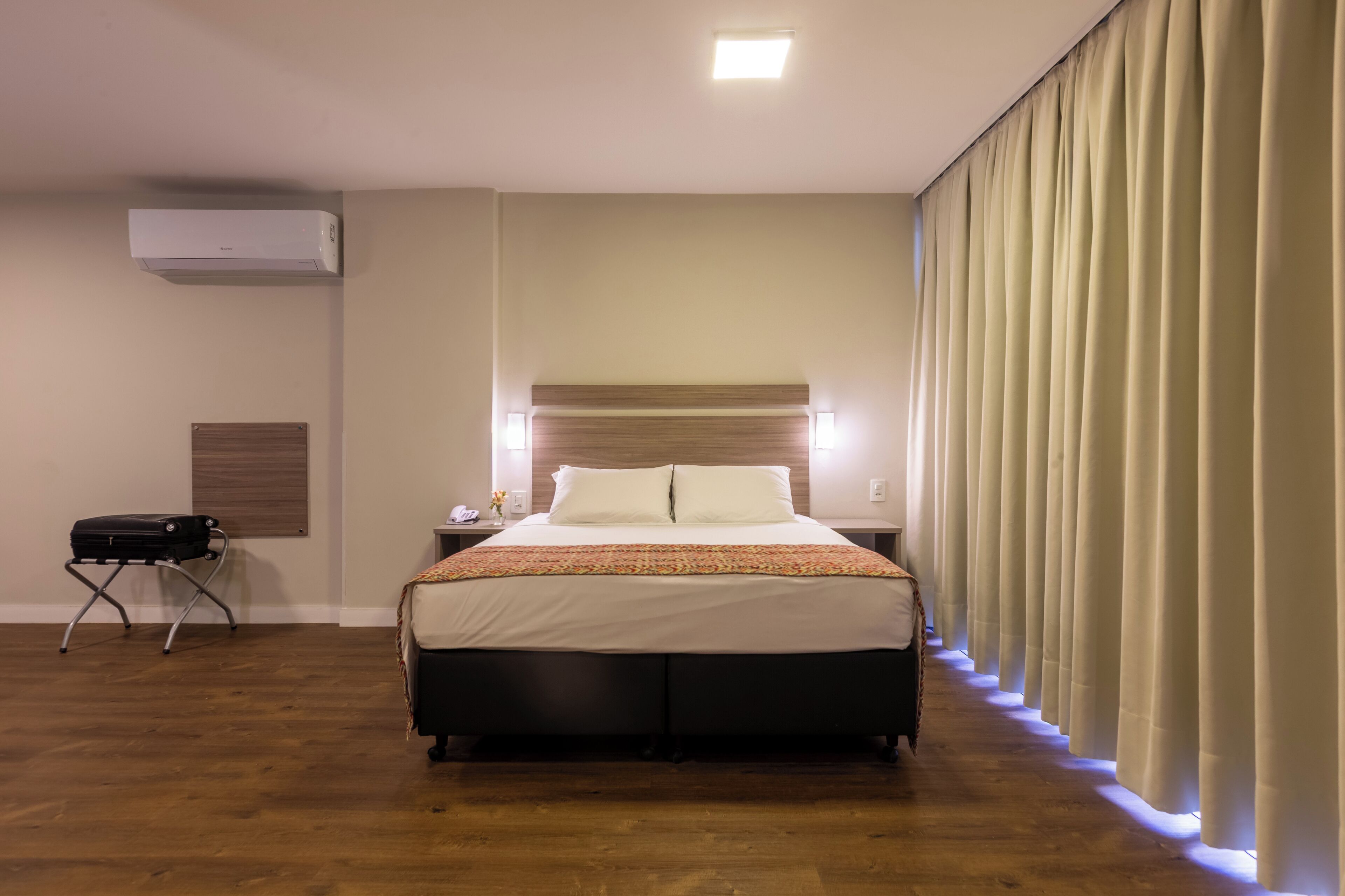 A hotel room with a bed, bedside lamps, a suitcase, an air conditioning unit, and a curtain.