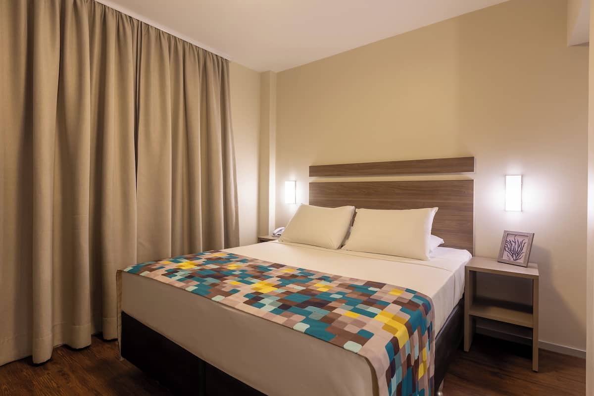 Une chambre d’hôtel avec un lit, une table de chevet, une lampe et une fenêtre avec des rideaux.