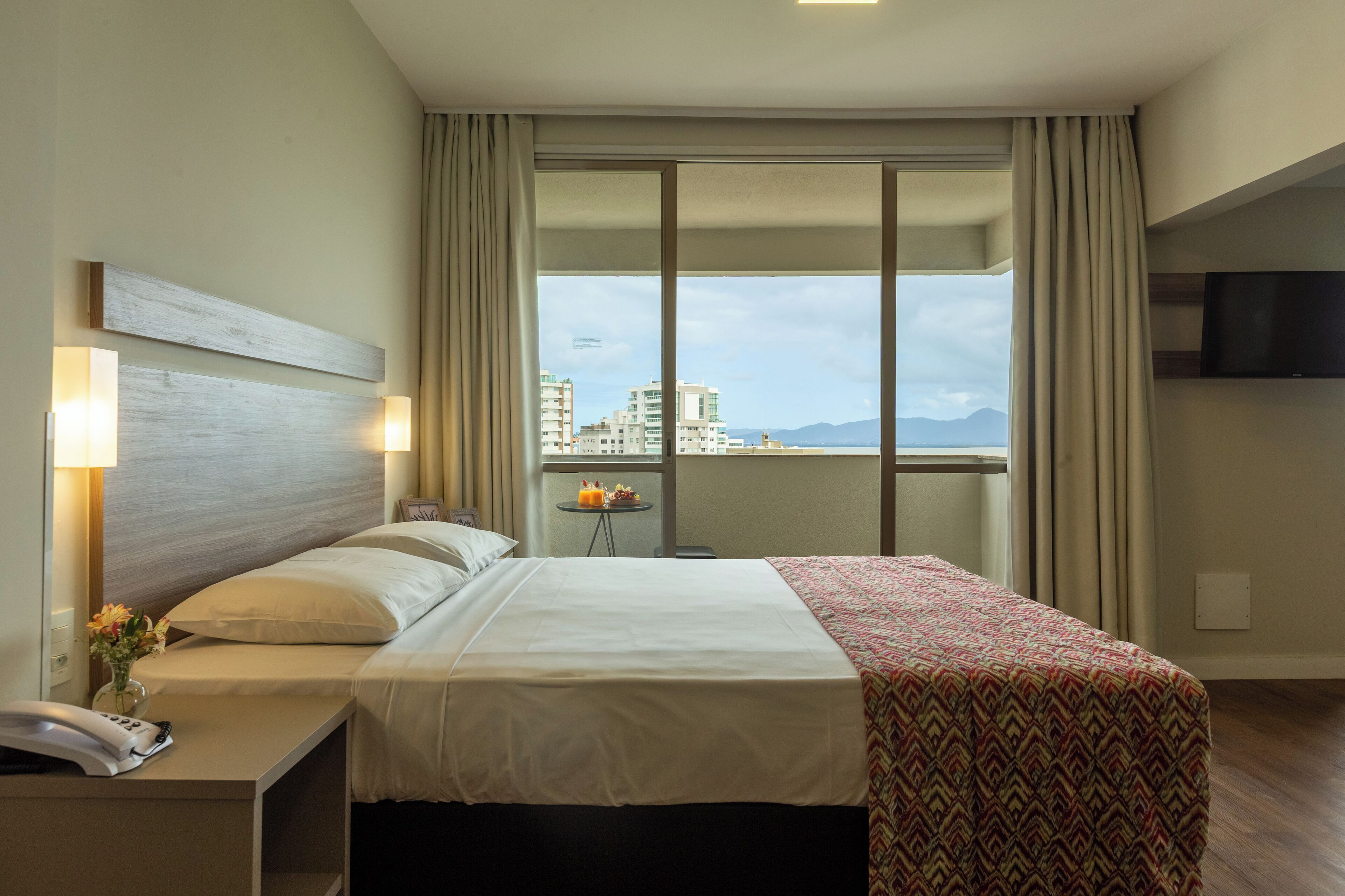 Una habitación de hotel con una cama grande, una mesita de noche con teléfono, una pequeña mesa con frutero y un televisor en la pared.
