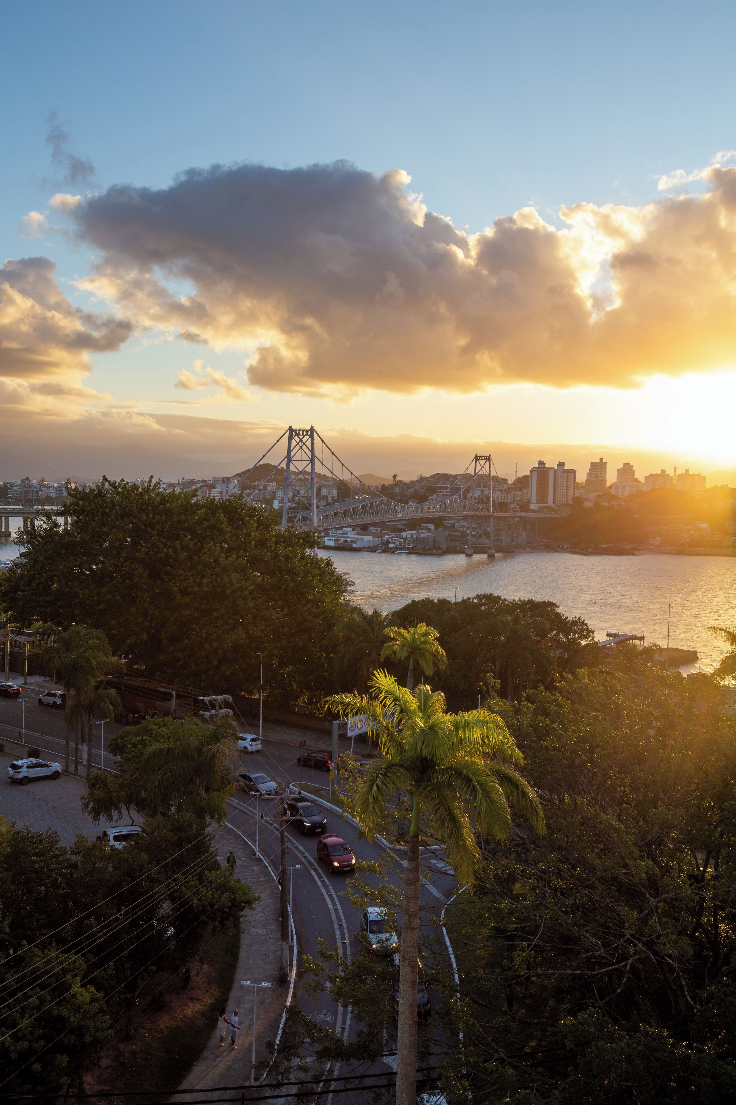 A sunset view of a city with a prominent bridge, a river, and surrounding greenery.