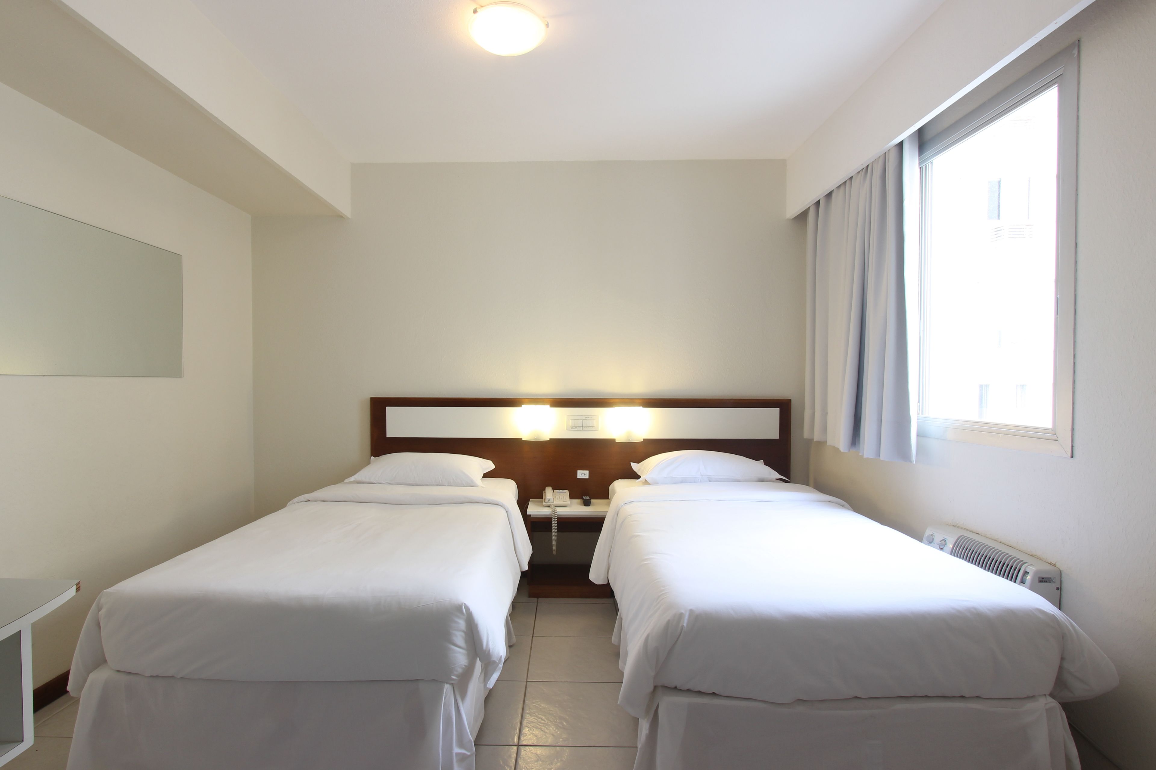A hotel room with two single beds, a bedside table with a lamp, a window with curtains, and a ceiling light.