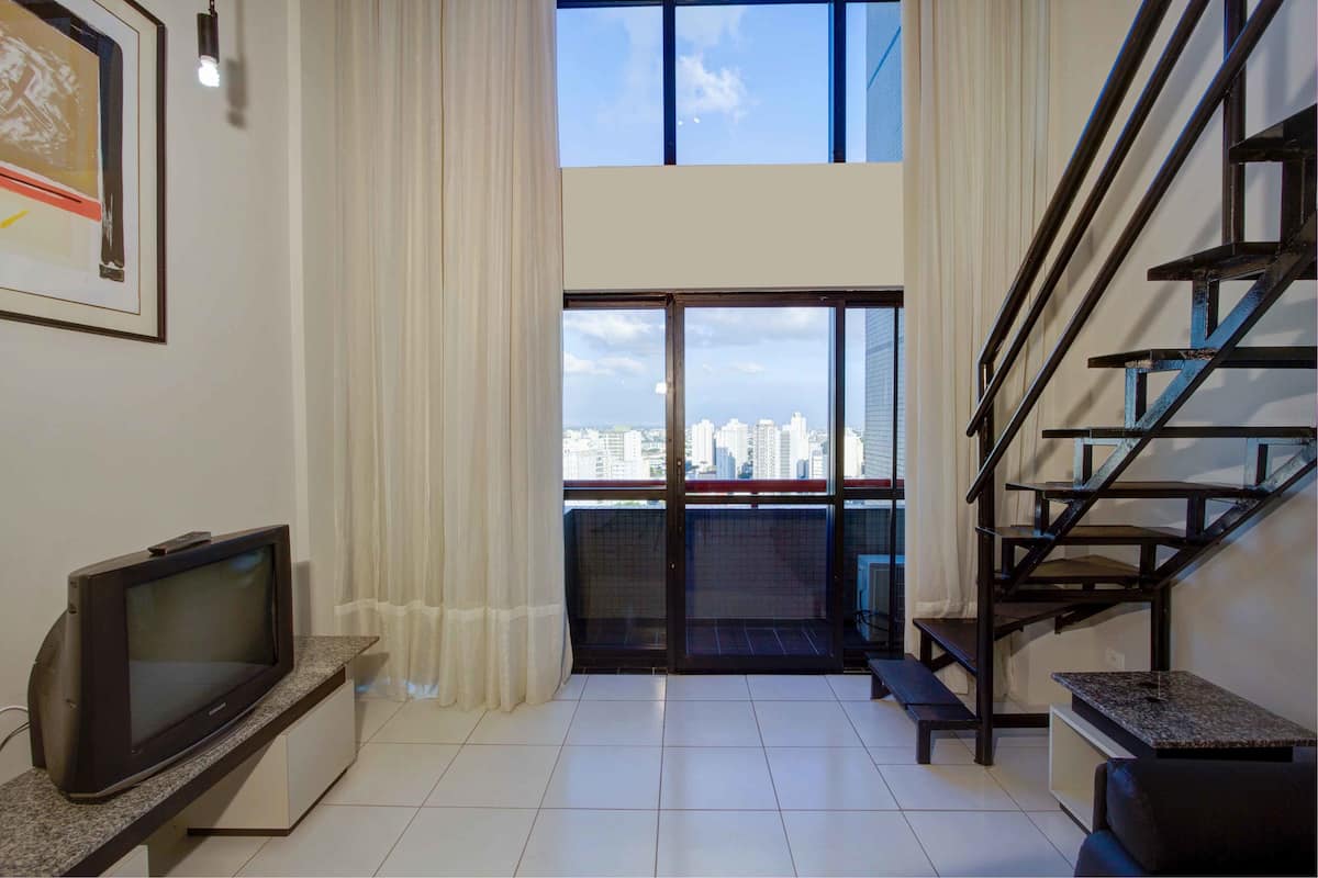 A modern living room with a television, a bench, and a staircase leading to a balcony with a city view.