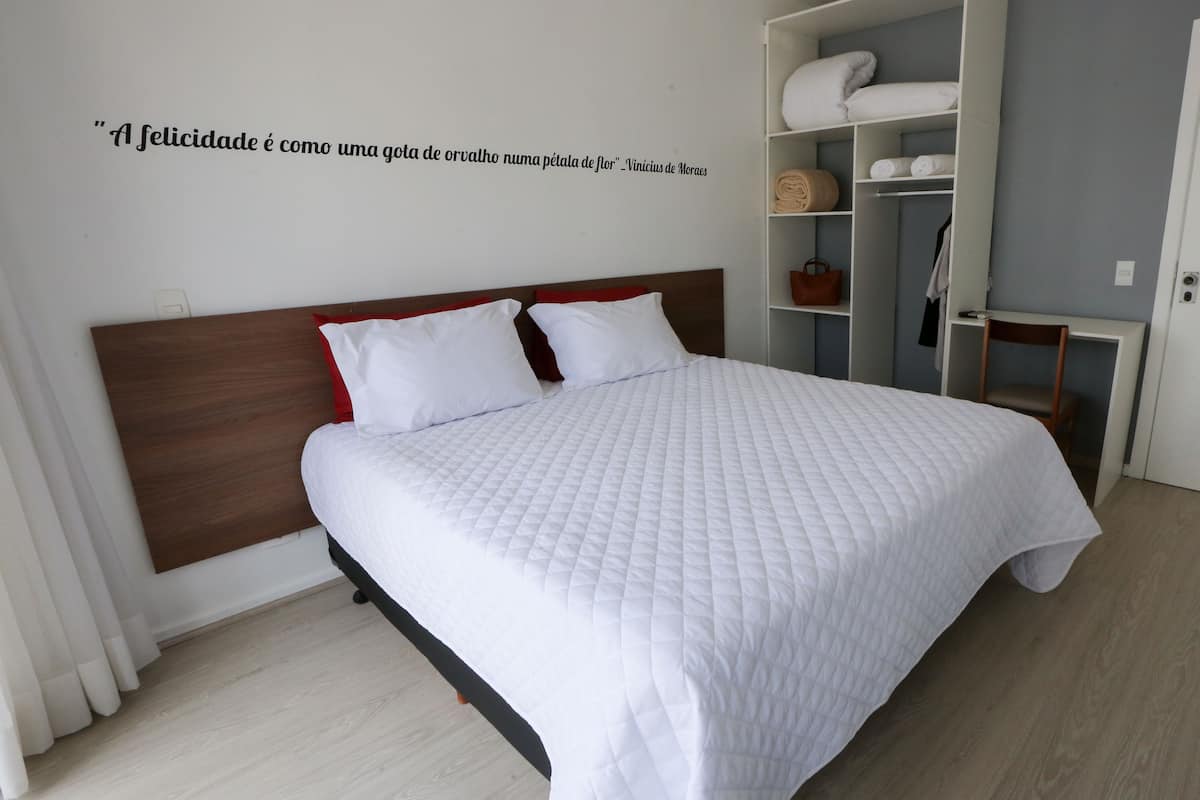 A neatly made bed with white bedding and red pillows, a wooden headboard, and a wall quote.