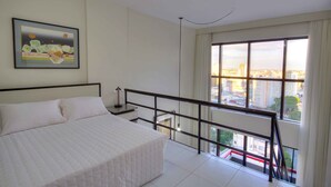 A bedroom with a large bed, a city view through a window, and a balcony with a railing.