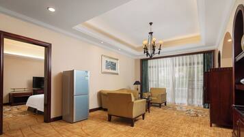 A hotel room with a bed, a refrigerator, two armchairs, a small table, a television, a chandelier, and a large window with curtains.