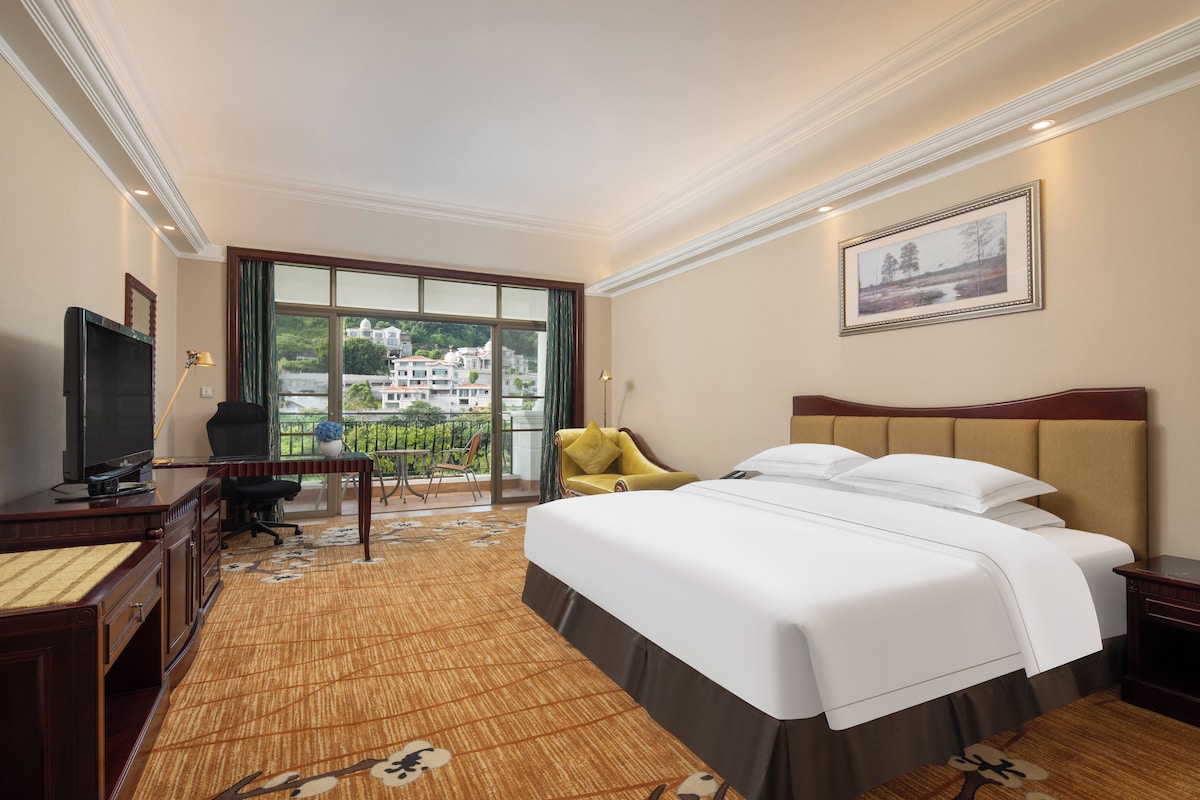 A hotel room with a large bed, a desk with a television, a chair, and a balcony with a view of buildings.